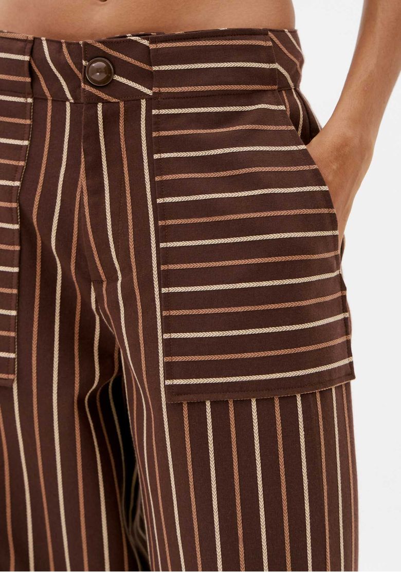 Close da calça barrel marrom listrada mostrando cintura média, botão frontal e bolso amplo com listras em direções contrastantes.