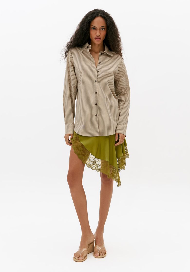 Camisa-feminina-oversized-listrada-bege-em-comprimento-alongado-combinada-com-saia-assimetrica-verde-e-sandalia-vista-de-corpo-inteiro